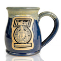 The front of the Bones Coffee Company Lucy hand thrown mug with the Atomic Apple art on the golden medallion. It is inspired by Zenimax and Amazon’s Fallout show. The mug is powder blue colored with a white and yellow glaze on top.