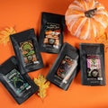 Five four ounce coffee bags are on an orange background with leaves and a pumpkin. They are named Apple Cider Donut, Frankenbones, Frosted Pumpkin Roll, Jacked o Lantern, and Blackberry Cobbler.