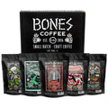 A box is behind five 12 ounce bags of flavored coffee. From left to right their names are Strawberry Cheesecake, Mint Invaders From Chocolate Space, White Russian, Maple Bacon, and Bananas Foster. The box features the Bones Coffee Company logo on it.